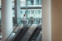 Rückansicht des Geschäftsmannes, der die Rolltreppe hinaufsteigt, während er im Büro auf sein Handy schaut — Stockfoto