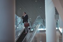 Vista baja del hombre de negocios con maletín hablando por teléfono en la escalera mecánica en la oficina - foto de stock