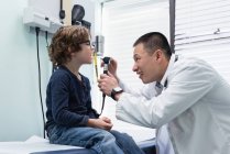 Visão lateral de jovem asiático médico masculino examinando caucasiano menino paciente com ferramenta em uma clínica — Fotografia de Stock
