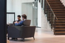 Vista lateral de empresários usando telefone celular sentado nas poltronas no lobby no escritório — Fotografia de Stock