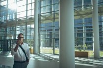 Vista frontal de un hombre de negocios hablando con el altavoz de su teléfono inteligente en la oficina contra la luz solar brillante - foto de stock