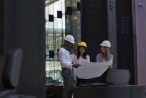 Вид сбоку группы архитекторов, обсуждающих проект в современном офисе. Они оснащены защитными шлемами — стоковое фото