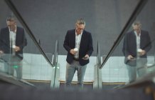 Blick auf Geschäftsmann mit Kaffeetasse, der sein Handy auf Rolltreppe im Büro benutzt — Stockfoto