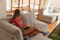 Visão de alto ângulo da mulher lendo livro enquanto se situa em casa na sala de estar — Fotografia de Stock