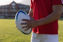 Sección media de un jugador de rugby masculino sosteniendo una pelota de rugby en el estadio en un día soleado - foto de stock