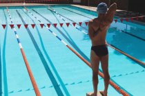 Vue arrière du jeune nageur masculin caucasien étirant les bras alors qu'il se tenait debout sur un bloc de démarreurs à la piscine extérieure le jour ensoleillé — Photo de stock