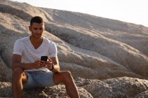 Vista frontal do homem afro-americano usando telefone celular perto do lado do mar enquanto sentado na rocha — Fotografia de Stock