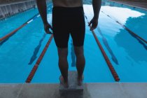 Section basse arrière d'un nageur mâle debout sur le bloc de départ devant la piscine — Photo de stock