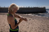 Vista lateral de una mujer mayor activa usando su reloj inteligente en la playa bajo el sol - foto de stock
