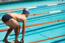 Visão traseira do jovem nadador caucasiano em pé na posição inicial no bloco de partida na piscina exterior no dia ensolarado — Fotografia de Stock