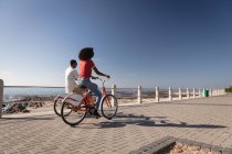 Vue arrière du couple afro-américain à vélo près de la mer par une journée ensoleillée — Photo de stock