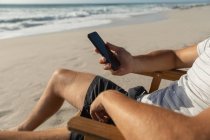 Розрив молодого чоловіка, який відпочиває на сонячному ліжку на пляжі в сонячний день. Він користується мобільним телефоном — стокове фото