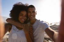 Vista frontal do casal afro-americano sorrindo e olhando para a câmera enquanto toma selfie perto do mar — Fotografia de Stock