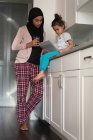 Vue à angle bas de mère mixte portant hijab et fille ensemble en utilisant une tablette numérique dans la salle de cuisine à la maison — Photo de stock