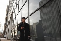Vorderansicht junger gut gekleideter asiatischer Geschäftsmann mit Handy, während er an modernen Gebäuden auf der Straße in der Stadt vorbeiläuft — Stockfoto