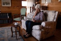 Frontansicht einer aktiven Seniorin, die ein digitales Tablet benutzt, während sie zu Hause auf dem Sofa sitzt und Tee trinkt — Stockfoto