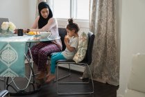 Side view of mixed race Mother wearing hijab talking with daughter while daughter eating orange fruit while sitting at table at home — Stock Photo
