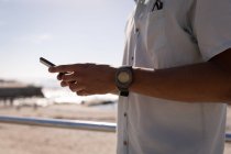 Seção média do homem de raça mista usando telefone celular na praia na luz do sol — Fotografia de Stock