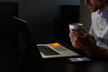 Seção intermediária de jovem executivo masculino tomando café enquanto trabalhava em laptop à mesa em um escritório moderno — Fotografia de Stock