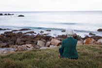 Задний вид вдумчивой активной пожилой женщины, сидящей на траве на пляже и смотрящей на море — стоковое фото