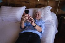 Високий кут зору сумної активної старшої жінки, яка використовує свій мобільний телефон, лежачи на ліжку в спальні вдома — стокове фото