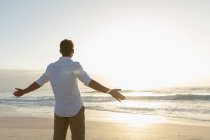 Вид сзади на расслабленного мужчину, стоящего на пляже в солнечный день. Он смотрит на закат в океане. — стоковое фото