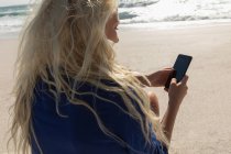 Вид збоку красива блондинка, використовуючи мобільний телефон на пляжі в сонячний день — стокове фото
