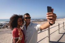 Vista frontal de la pareja afroamericana de pie y hablando selfie mientras sonríe en la playa en un día soleado - foto de stock