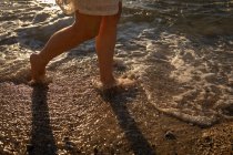 Unterteil einer aktiven Seniorin, die am Abend am Ufer des Strandes spaziert, während sich der Sonnenuntergang im Wasser spiegelt — Stockfoto