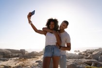 Vista frontal do casal afro-americano de pé e tomando selfie perto do lado do mar na rocha — Fotografia de Stock