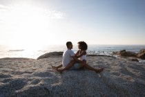 Side view of African-american couple in romantic mood sitting on rock near sea side and looking at each other — стоковое фото