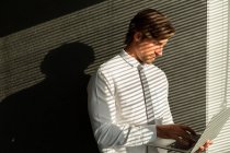 Vista frontale di bel giovane dirigente di sesso maschile che lavora sul computer portatile mentre in piedi vicino alla finestra in un ufficio moderno — Foto stock