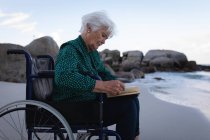 Vue latérale d'une aînée active handicapée lisant un livre sur un fauteuil roulant près de l'eau sur la plage — Photo de stock