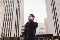 Front view of young Asian businessman with coffee cup talking on mobile phone in city surrounded by tall business buildings — Stock Photo