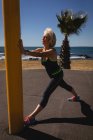 Vista laterale di una donna anziana attiva che si estende contro un palo vicino alla spiaggia sotto il sole — Foto stock