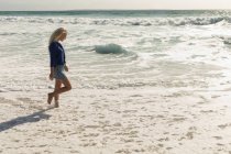 Vista lateral de la mujer rubia caminando en la playa en un día soleado. Ella está caminando. - foto de stock