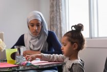 Вид спереду змішаної скаченої жінки в хіджабі і дочка розмовляє один з одним вдома, сидячи на стільці навколо столу — стокове фото