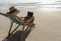 Vista trasera de la joven mujer que se relaja en la tumbona en la playa en un día soleado. Ella está usando su portátil - foto de stock