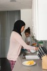 Vista lateral de la madre de raza mixta con hijab lavabo debajo del fregadero, mientras que la hija mirando fregadero en la cocina en casa - foto de stock