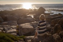 Vista posteriore di una donna anziana attiva che si rilassa sulla roccia e guarda il tramonto sulla spiaggia — Foto stock