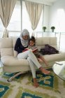 Vista frontal da mãe de raça mista vestindo hijab e filha sentado e lendo livro na sala de estar em casa — Fotografia de Stock