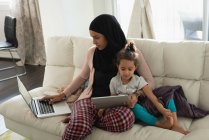Вид спереди на смешанную расу Мать носит хиджаб, используя ноутбук, в то время как дочь смотрит на цифровой стол в гостиной дома — стоковое фото