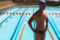 Vorderansicht junger kaukasischer Schwimmer, der mit den Händen auf der Hüfte steht, während er Badebekleidung trägt und am sonnigen Tag im Freibad zusieht — Stockfoto