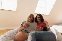 Низкий угол обзора разнообразных женщин, улыбающихся и пользующихся цифровым планшетом дома на диване в спальне — стоковое фото