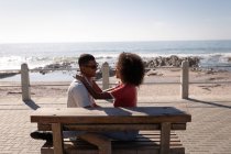 Vista lateral do casal afro-americano de humor romântico sentado na prancha de madeira perto do lado do mar. Eles estão sentados cara a cara enquanto sorriem — Fotografia de Stock