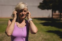 Frontansicht einer aktiven Seniorin, die ihre Kopfhörer trägt, um Musik zu hören, während sie an einem sonnigen Tag im Park trainiert — Stockfoto