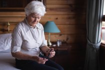 Вид спереди активной пожилой женщины, проверяющей уровень сахара в крови глюкометром на кровати в спальне дома — стоковое фото