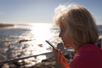 Крупный план активной пожилой женщины, разговаривающей по мобильному телефону на набережной перед морем в лучах солнца — стоковое фото