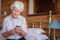 Vista frontal de una mujer mayor activa sentada en la cama y usando su teléfono móvil en el dormitorio en casa - foto de stock