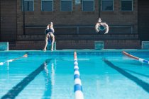 Vista frontal de nadadores caucásicos masculinos y femeninos saltando al agua al mismo tiempo en la piscina bajo el sol - foto de stock